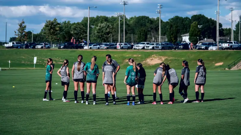 A girls' team in grey and teal training kit gathered together on a wide sunlit field