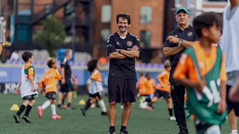 Two coaches watching a session of young players in orange and green bibs at Truist Point
