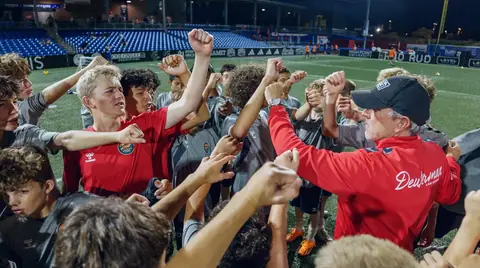 Players and a coach with their fists raised together in a huddle after a floodlit session