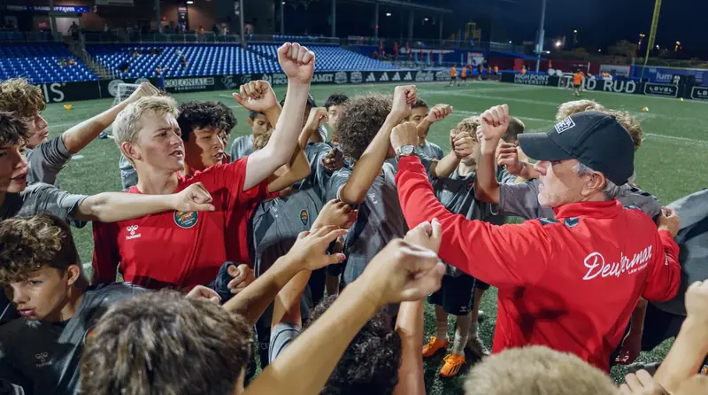 Players and a coach with their fists raised together in a huddle after a floodlit session
