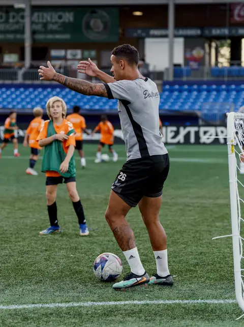 A Carolina Core first-team player, arms outstretched, coaching a group of young players in orange and green bibs beside a small goal