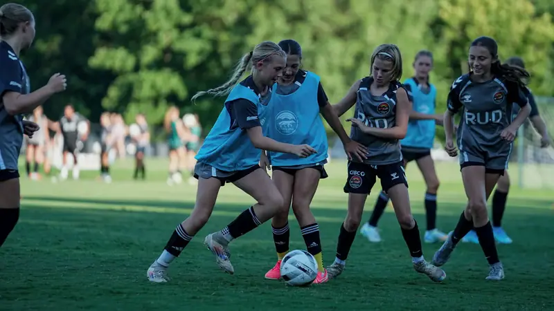 A Carolina Core first-team player, arms outstretched, coaching a group of young players in orange and green bibs beside a small goal