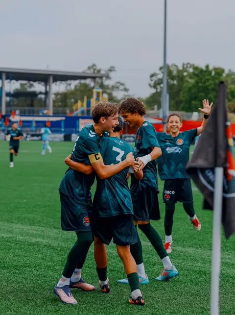 Two academy players in teal kit celebrating a goal with their arms around each other as a team-mate runs in