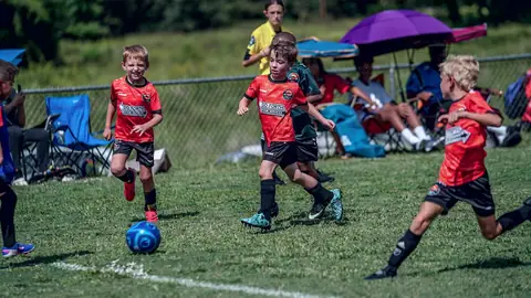 Three U7 players in Orange CCFC Kits chase after the ball at Bagley Sports Complex