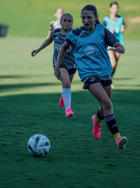 A young player in a light-blue bib on the ball at a Pre-GA training session, a teammate closing in behind