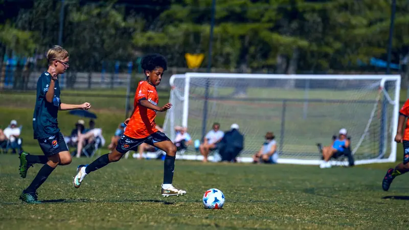 Two CCFC U11 players chase after the ball, One in an Orange CCFC Kit and one in a Green CCFC kit with parents watching in the background