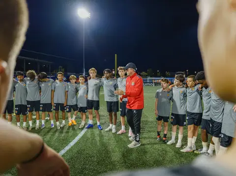 A coach in a red jacket talking to a circle of academy players under the floodlights, seen over a player's shoulder