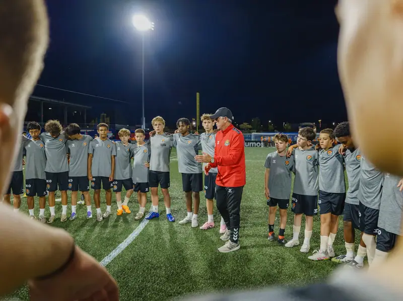 A coach in a red jacket talking to a circle of academy players under the floodlights, seen over a player's shoulder