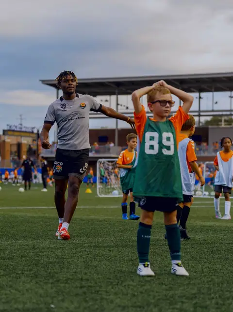 A young player in a green bib grinning with his hands on his head as a first-team player walks past